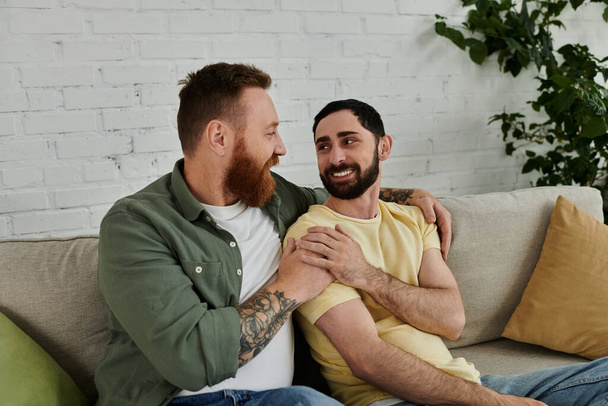 Ένα ζευγάρι γενειοφόρων ανδρών χαλαρώστε και να συνομιλήσετε πάνω από έναν καναπέ σε ένα άνετο σαλόνι. - Φωτογραφία, εικόνα