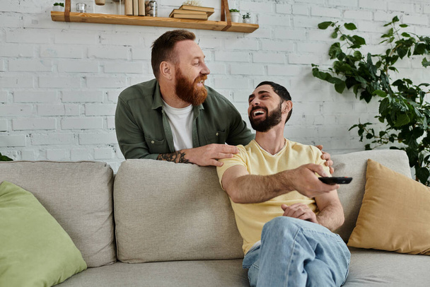 Een bebaarde gay paar zit op een bank, het vasthouden van een afstandsbediening, televisie kijken - Foto, afbeelding