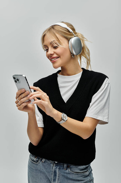 Egy fiatal szőke nő elegáns öltözékben fülhallgatót visel, és mobiltelefont használ szürke alapon.. - Fotó, kép