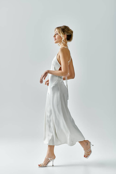 Витончена молода жінка з світлим волоссям у білій сукні елегантно ходить на сірому фоні. - Фото, зображення