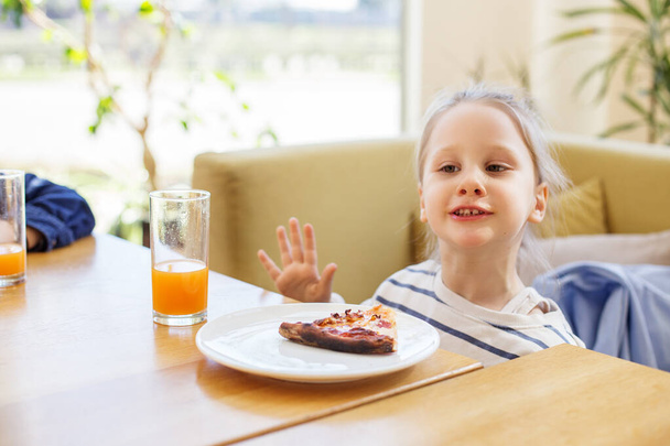 Alegre niño saludando a la cámara mientras disfruta de una rebanada de pizza y un vaso de jugo de naranja. - Foto, imagen