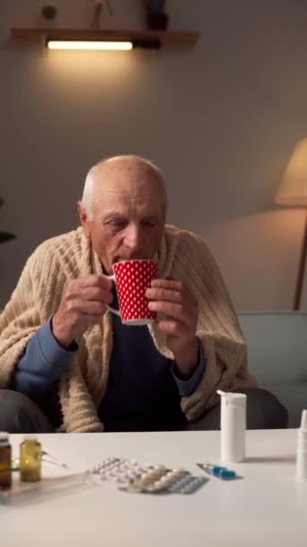 L'uomo anziano si congela in inverno avvolto in plaid, tenendo in mano una tazza di bevanda calda a casa che soffre di febbre. Video verticale - Filmati, video