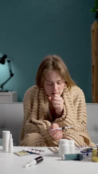 Een vrouw gewikkeld in een deken controleert haar temperatuur met een thermometer en heeft medicijnen op tafel. - Video
