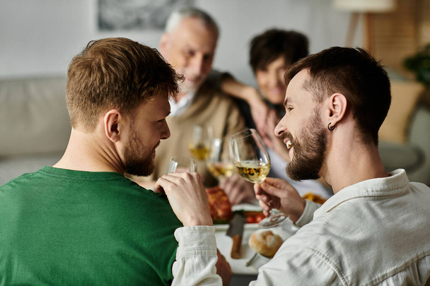 Ένα γκέι ζευγάρι απολαμβάνει ένα δείπνο με τους γονείς του στο σπίτι, μοιράζοντας μια πρόποση. - Φωτογραφία, εικόνα