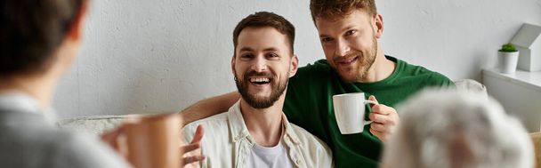 Um casal gay sorri e segura canecas enquanto está sentado em um sofá em sua casa, provavelmente se encontrando com a família. - Foto, Imagem