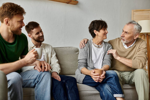 Ένα γκέι ζευγάρι κάθεται με τους γονείς του σε έναν καναπέ, συμμετέχοντας σε μια ζεστή και οικεία συζήτηση. - Φωτογραφία, εικόνα
