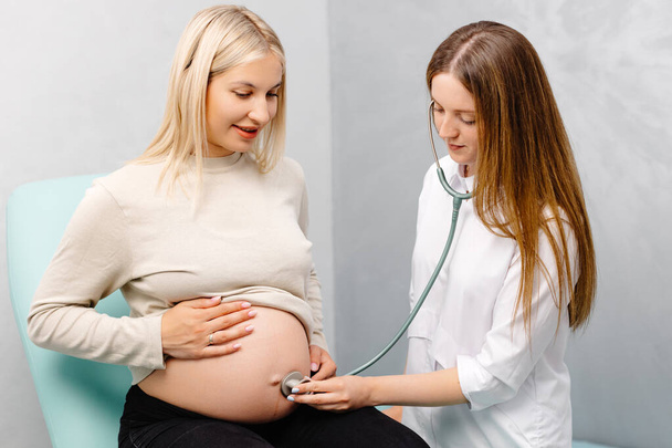 Жінка-лікар, яка використовує стетоскоп, вивчає вагітну жінку в клініці. Медичний огляд вагітної жінки
. - Фото, зображення