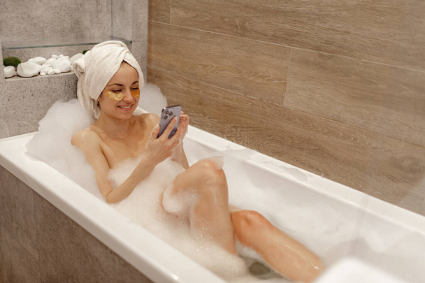 Usmívající se sexy hezká mladá žena s páskou přes oko a ručníkem na hlavě, jak píše do telefonu, zatímco leží ve vaně s pěnou. Hygiena a koncepce mytí těla. Technologie, komunikace - Fotografie, Obrázek