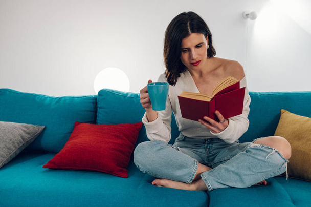 Junge entspannte und schöne kaukasische Frau in lässigem Outfit liest ein Buch und trinkt Tee, während sie es sich auf einem Sofa in einem gemütlichen Wohnzimmer zu Hause gemütlich macht. - Foto, Bild