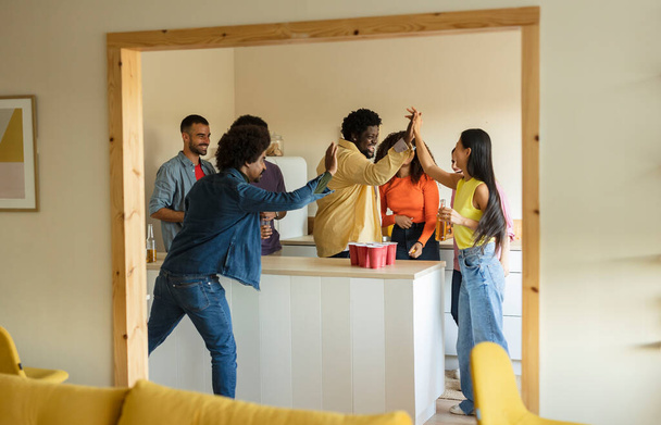 Groupe d'amis dans la vingtaine et la trentaine jouant au bière-pong, célébrant avec des hauts-cinq et des boissons dans un cadre confortable à la maison. - Photo, image