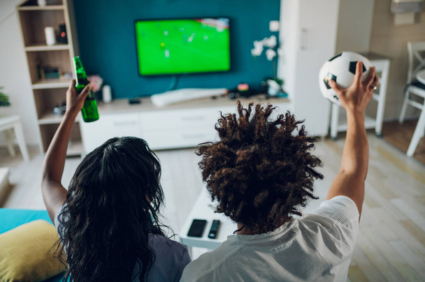 Afroamerikanisches Sportfan-Paar schaut sich wichtige Fußballspiele im Fernsehen an, während es zu Hause Bier trinkt und jubelt. Guy hält einen Fußball in der Hand. Jubel bei einer Lieblingsmannschaft. Rückansicht. - Foto, Bild