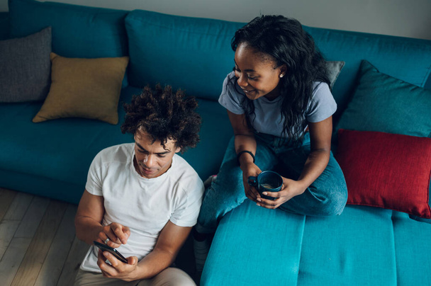 明るいアフリカ系アメリカ人のカップルは,コーヒーを飲みながらスマートフォンを使用し,週末のリビングルームで時間を過ごしながら会話をする.. - 写真・画像
