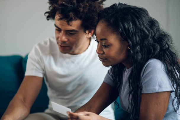 Portrait of a young african american couple holding a credit card while managing their expenses at home and calculating their home budget. Focus on the woman. - Photo, Image