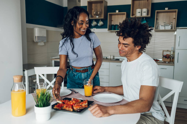 Šťastný milennial africký americký pár těší zdravé snídaně, zatímco sedí u kuchyňského stolu doma. Jím toasty a piju pomerančový džus. Směje se a mluví. - Fotografie, Obrázek