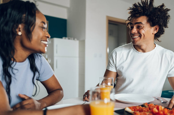 ハッピーミレニアルアフリカ系アメリカ人のカップルは,自宅のキッチンデスクに座って健康的な朝食を楽しんでいます. トーストとオレンジジュースを食べる. 笑って話す. - 写真・画像