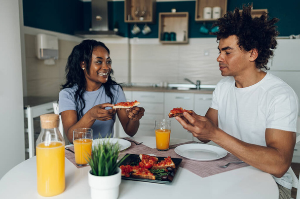 Glückliches junges afrikanisch-amerikanisches Paar, das Spaß in der Küche hat, gesundes Essen isst und Orangensaft trinkt.. - Foto, Bild