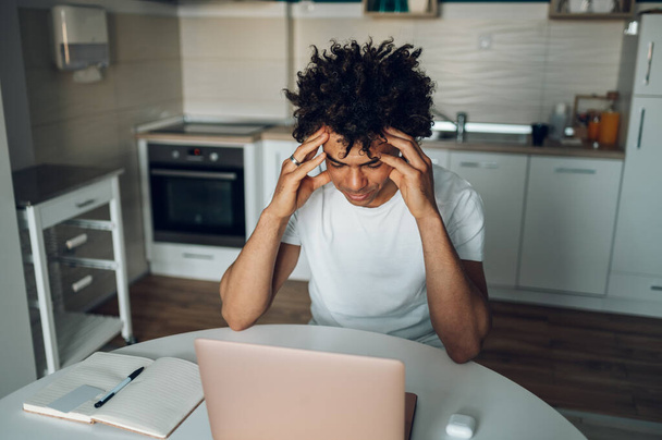 Втомлений молодий афроамериканський чоловік відчуває стрес, працюючи на ноутбуці і сидячи на кухні вдома. Робота і термін. Проблеми. - Фото, зображення