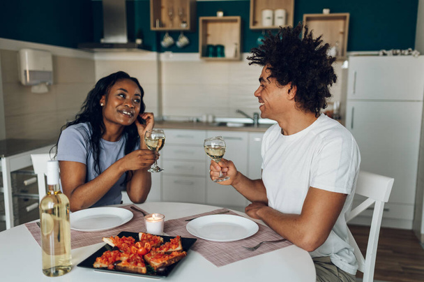 Romantisches afrikanisch-amerikanisches Paar, das zu Hause in der Küche Weißwein trinkt und zum Abendessen eine gesunde Mahlzeit zu sich nimmt. Wochenende zu Hause. - Foto, Bild