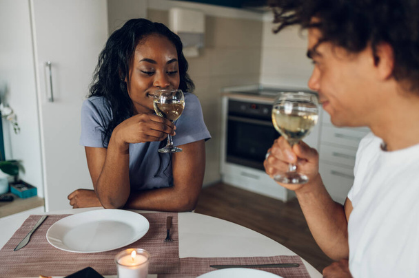 Веселая африканская американская пара ужинает вместе дома и празднует свой особенный день, выпивая белое вино. Романтическая пара празднует годовщину. - Фото, изображение