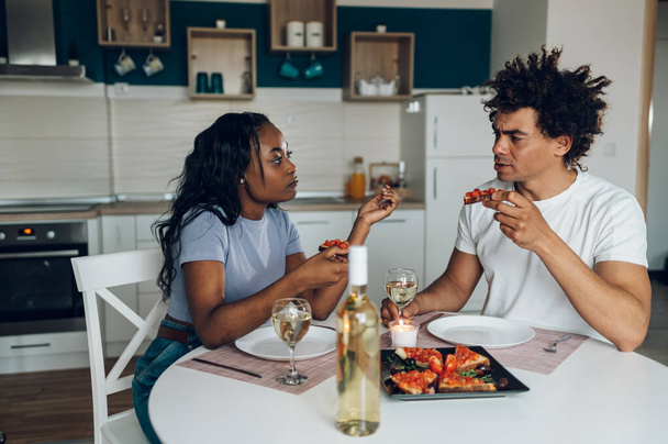 Wesoła afrykańska para jadła razem w domu i świętowała swój wyjątkowy dzień pijąc białe wino. Romantyczna para świętująca rocznicę. - Zdjęcie, obraz