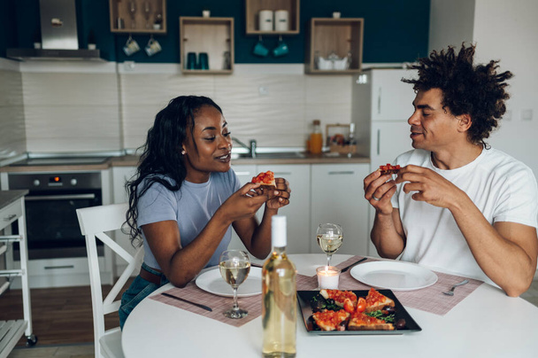 Romántica pareja afroamericana brindando bebiendo vino blanco en la cocina en casa mientras come una comida saludable para la cena. Fin de semana en casa. - Foto, imagen