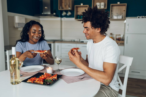 Allegra coppia afroamericana che cena insieme a casa e celebra il loro giorno speciale bevendo vino bianco. Coppia romantica che celebra anniversario. - Foto, immagini