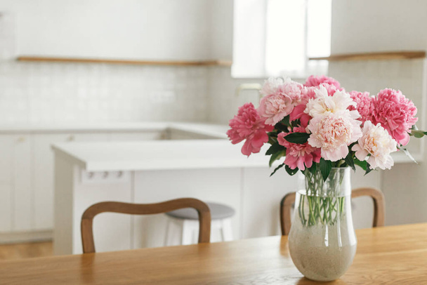 Prachtige pioenrozen in vaas op houten tafel op de achtergrond van stijlvolle witte keuken met eiland, houten planken en apparatuur in nieuw huis. Moderne keuken interieur in boerderij - Foto, afbeelding