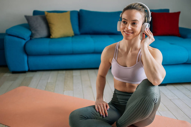 Mujer deportiva joven sentada en una esterilla de yoga y escuchando música en los auriculares y usando un teléfono inteligente mientras se entrena en casa. Practicando meditación. Disfrutando de su lista de reproducción favorita. - Foto, Imagen