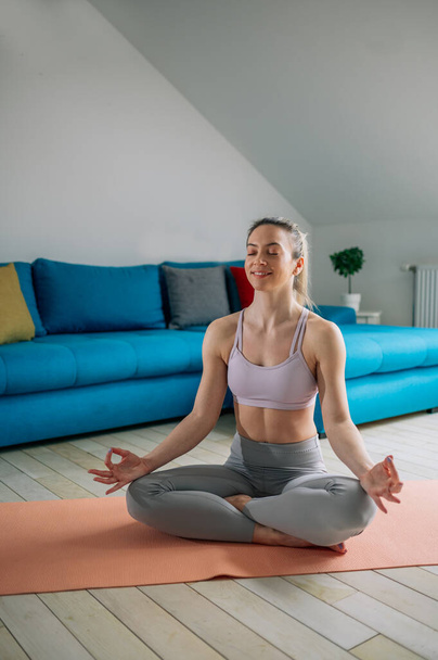 Atraktivní mladá žena sedící v pozici lotosové jógy na domácí fitness podložce v obývacím pokoji. Uvolňující stav mysli, meditace. Žádný stresový koncept. - Fotografie, Obrázek