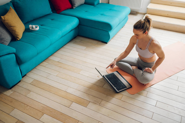 Genç atletik kadın evde antrenman yapıyor ve bir dizüstü bilgisayarla çevrimiçi antrenman izlerken egzersizleri tekrarlıyor. Sağlıklı yaşam tarzı. - Fotoğraf, Görsel