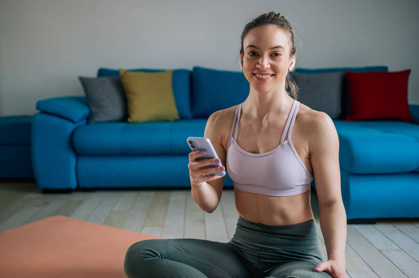 Beautiful athletic woman using airpods and a smartphone while listening music and relaxing after training at home. Using smartphone during workout break. - Photo, Image