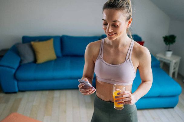 Mujer atlética joven bebiendo jugo de naranja después de entrenar en casa y usando un teléfono inteligente. Estilo de vida saludable. - Foto, Imagen