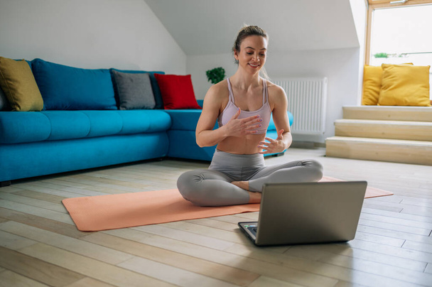 Χαρούμενη δραστήρια γυναίκα κάνει fitness στο σπίτι, ενώ χρησιμοποιεί φορητό υπολογιστή για να παρακολουθήσετε online μαθήματα. Βλέποντας βίντεο γυμναστικής στο διαδίκτυο. Μάθημα βίντεο. - Φωτογραφία, εικόνα