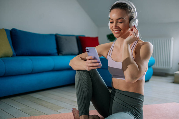 Sportos fiatal nő hallgat kedvenc zene fülhallgató és ellenőrzése fitness app vagy a szociális hálózatok okostelefon edzés után otthon. Sport, technológia és egészséges életmód koncepció. - Fotó, kép