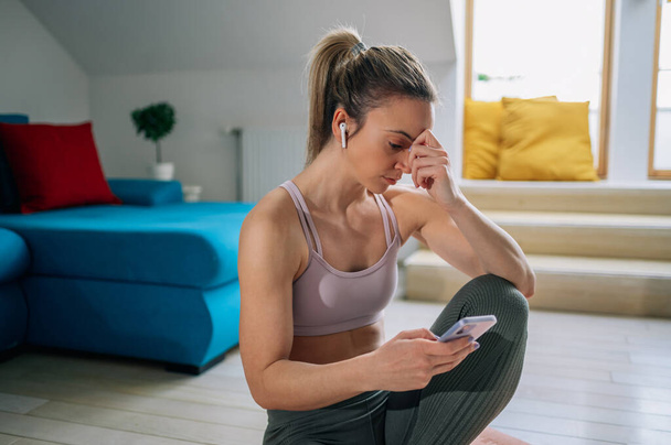 Young sporty woman sitting on a yoga mat and listening music on a airpods and using a smartphone while training at home. Practicing meditation. Enjoying her favorite playlist. - Photo, Image