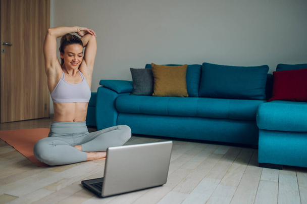 Bella giovane donna che si allena a casa e si allunga il corpo sul tappetino yoga mentre guarda il tutorial sul computer portatile in soggiorno. Esercizio a casa. - Foto, immagini