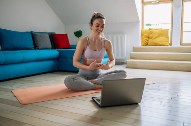Молодая женщина тренируется дома и растягивает тело на коврике для йоги, наблюдая за уроком на ноутбуке в гостиной. Упражнения на дому. - Фото, изображение