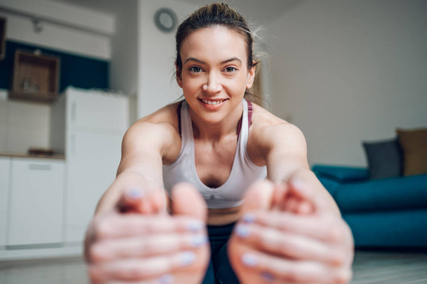 Mujer atlética extendiéndose para tocarse los dedos de los pies mientras está sentada en una esterilla de yoga y entrenando en casa en el suelo. Estirar antes de hacer ejercicio. Planta de flexión hacia adelante sentada. - Foto, imagen