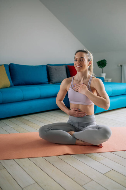 Joven mujer hermosa sana en top deportivo y polainas practicando yoga en casa mientras está sentada en pose de loto sobre esterilla de yoga y meditando. Concepto de meditación Mindfulness. - Foto, imagen