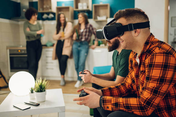 Male best friends having fun with virtual reality headset glasses while playing online video games and spending time together at home. Female friends blurry in the background. - Photo, Image