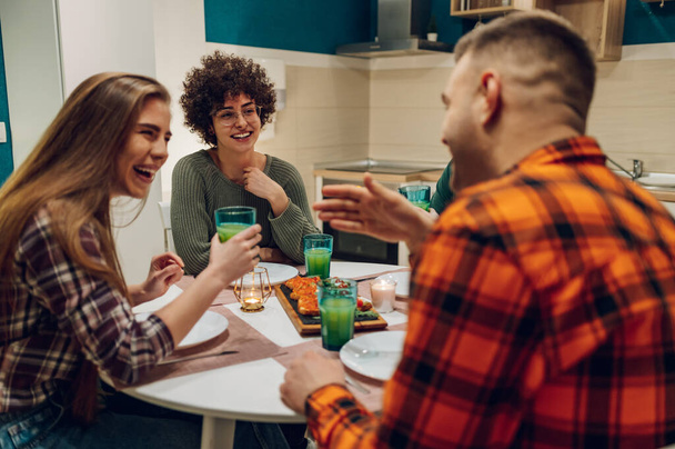 Fröhliche junge Leute genießen das Essen, während sie gemeinsam am Esstisch in der Küche sitzen. Freunde treffen sich zu Hause zum Abendessen. - Foto, Bild