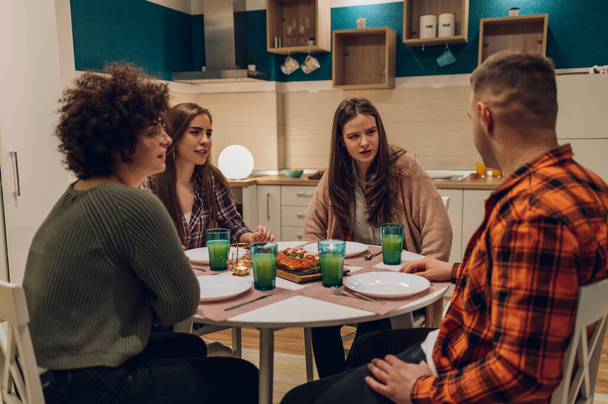 Barátok ülnek a konyhaasztalnál és beszélgetnek vacsora közben. Fiatal boldog emberek kommunikálnak, miközben együtt vacsoráznak otthon az asztalnál.. - Fotó, kép