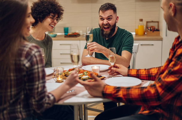 Grupa wesołych młodych ludzi korzystających z kolacji, siedząc razem przy stole w kuchni. Świetnie się bawisz z przyjaciółmi podczas kolacji podczas picia i zabawy - Zdjęcie, obraz