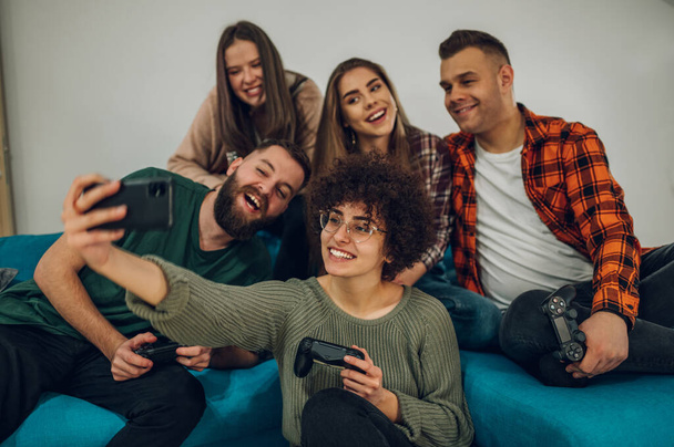 Χαρούμενη ομάδα φίλων που διασκεδάζουν στο σπίτι και βγάζουν selfie με ένα smartphone ενώ κάθονται στον καναπέ. Έννοια φιλίας και τεχνολογίας. - Φωτογραφία, εικόνα