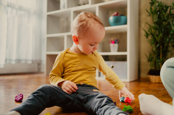 Portret dziecka bawiącego się kolorową gliną siedzącego na podłodze. Dzieci się bawią. Mały chłopiec tworząc obiekty i kształty podczas formowania gliny lub plastycyny. Gra edukacyjna dla niemowląt i małych dzieci. - Zdjęcie, obraz
