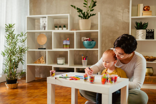 Πορτρέτο της μητέρας και του παιδιού γιο παίζει και ζωγραφίζει μαζί στο μικρό τραπέζι στο σπίτι. Babysitter γυναίκα ή θεία βοηθώντας το παιδί αγόρι, ενώ έχει τη διασκέδαση. Ευτυχισμένη οικογένεια. Αντιγραφή χώρου. - Φωτογραφία, εικόνα