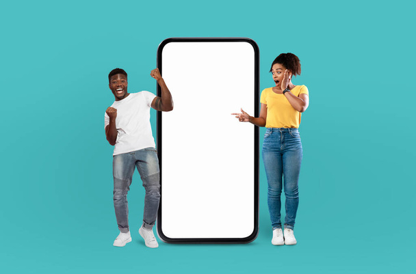 Casal afro-americano em pé ao lado de uma tela de telefone em branco, olhando para ele atentamente. A tela é desprovida de qualquer conteúdo, exibindo apenas uma superfície reflexiva. - Foto, Imagem