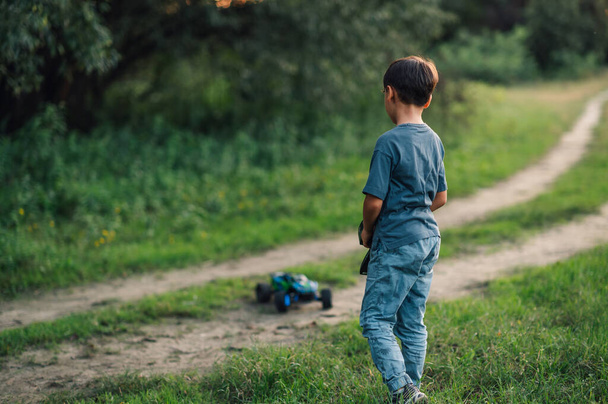 Фотография мальчика, наблюдающего за своей игрушечной машиной, бегущей по грунтовой дороге на природе. Он управляет им с помощью дистанционного управления. Размытый фон с сельскими пейзажами, травой и деревьями. - Фото, изображение