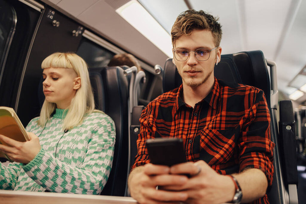Portrét dojíždějícího cestujícího vlakem, který si přivazuje mobil, zatímco se na něj dívá během cesty. Vedle něj je dívka s knihou. Mladý hipster cestovatel používá svůj telefon ve vlaku. - Fotografie, Obrázek