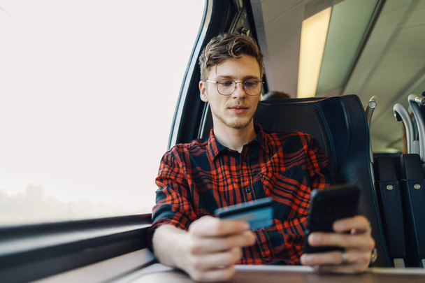 Portrét mladého cestovatele sedícího ve vlaku s kreditní kartou a mobilem v rukou a kupujícího si jízdenku na vlak online v mobilní aplikaci. Dojíždějící používá aplikaci e-banking ve vlaku během své cesty. - Fotografie, Obrázek